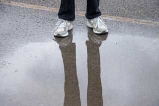 A person standing in the middle of a street with their reflection in a puddle
